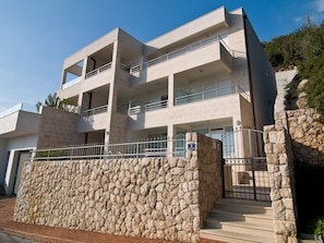 A modern multi-story building with balconies, a stone wall, and a gated entrance.
