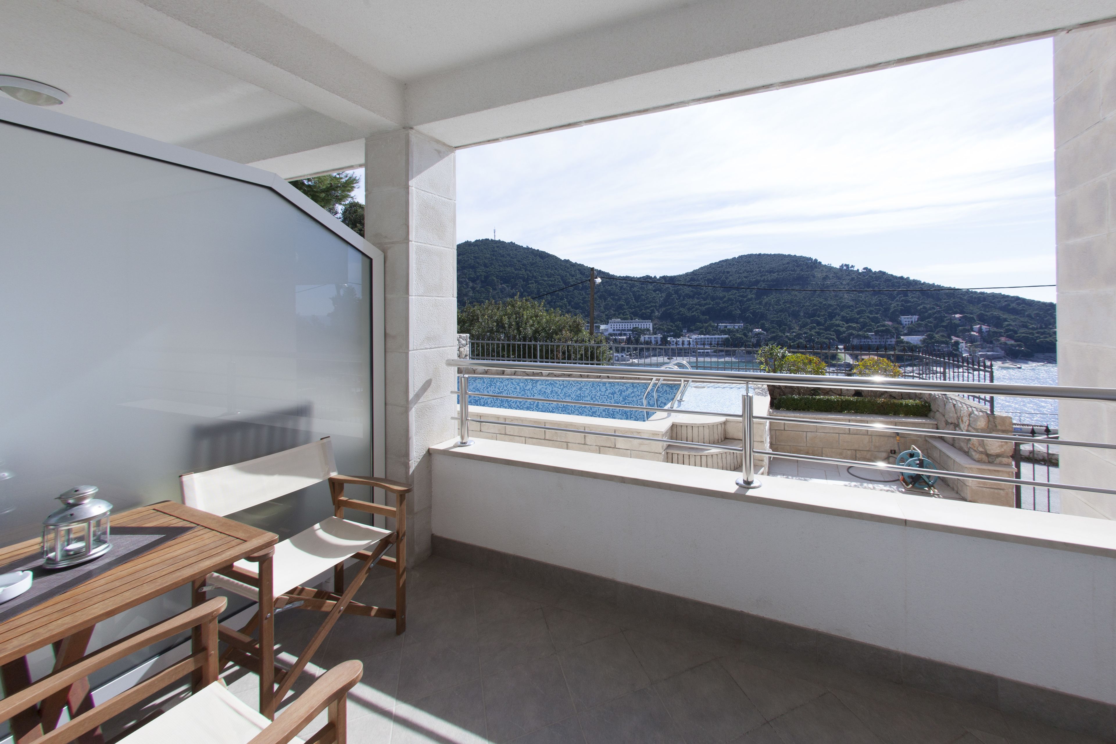 A balcony with a view of a body of water and hills, a table with chairs, and a railing.