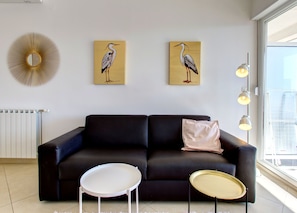 Uma sala de estar moderna com um sofá preto, duas pinturas de pássaros, uma mesa redonda branca e uma mesa redonda pequena.