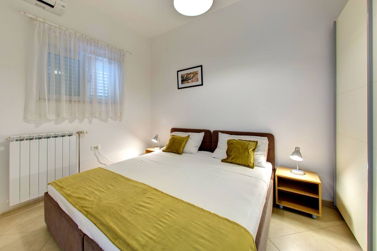 Un dormitorio con cama, mesitas de noche, radiador, ventana con cortinas traslúcidas y un cuadro en la pared.