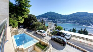 Una piscina con vistas al mar y a las montañas, un estacionamiento con coches y un muro de piedra.