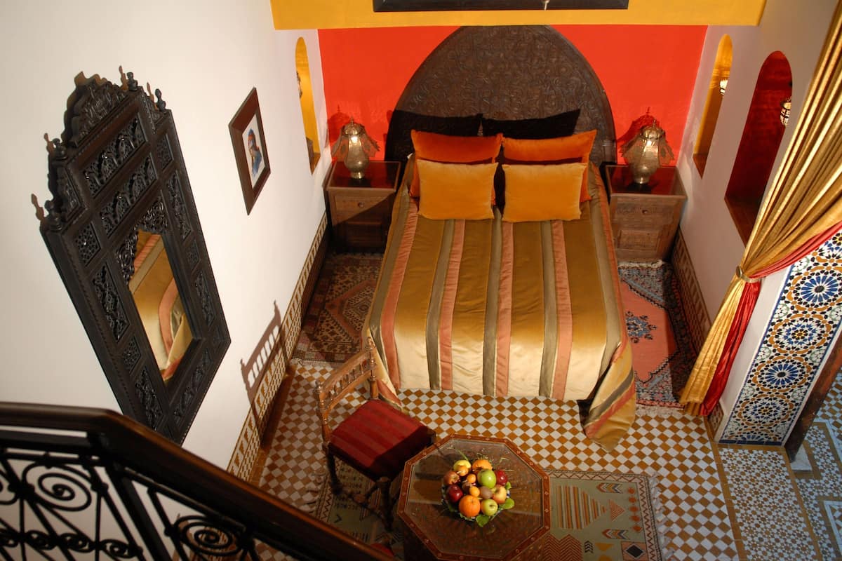 A room with a checkered floor, a bed with striped bedding, a wooden chair, a small table with fruit, and a decorative mirror.