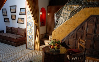 A room with a checkered floor, a sofa, a table with fruit, a staircase with a metal railing, and patterned walls.