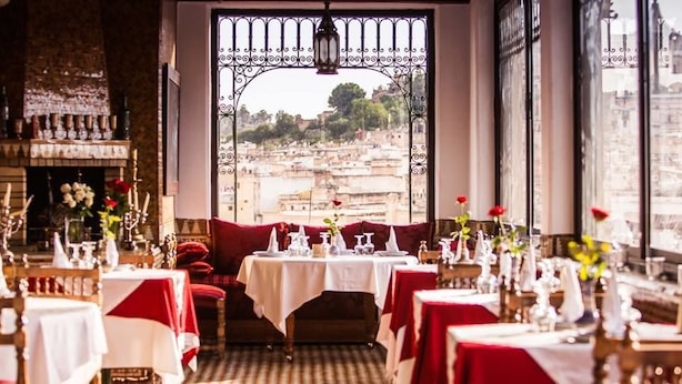 A restaurant with tables set for dining, a fireplace, and a view of a cityscape through a large window. - Dar Tazi - Medina View (Fes)