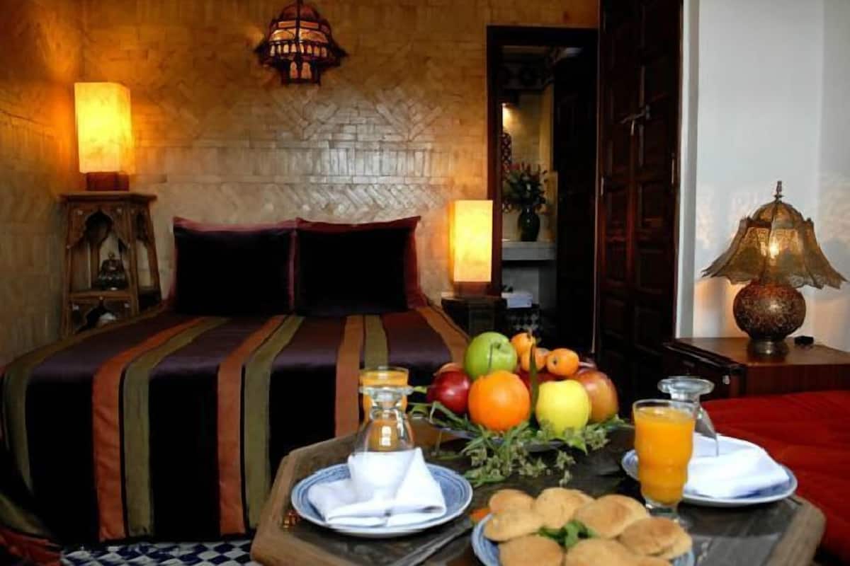 A neatly made bed with a striped bedspread, a bedside table with a fruit bowl, a glass of orange juice, and a bottle of water.
