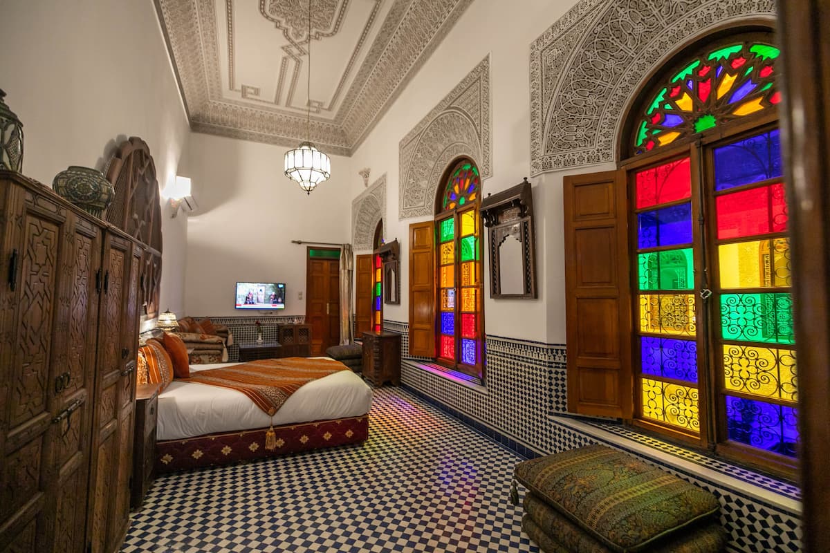 A room with a checkered floor, a bed with a white and red bedspread, wooden furniture, and colorful stained glass windows.