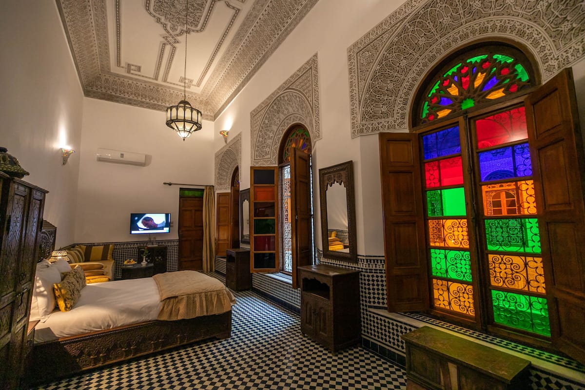 A bedroom with a checkered floor, a bed, a television, and a stained glass window.