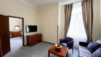 A hotel room with a sofa, a television, a wooden cabinet, a small table with fruit, and a window with curtains.