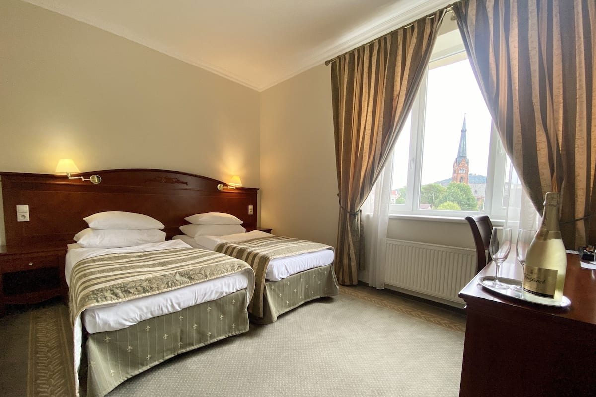 A hotel room with two beds, a wooden headboard, a window with curtains, a radiator, and a table with a bottle and glasses.