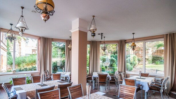 Een restaurant met meerdere tafels en stoelen, grote ramen met gordijnen en hangende kroonluchters.