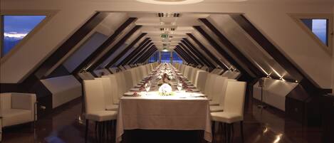 A long dining table with white chairs, set for a formal meal, in a modern interior with a high ceiling and skylights.