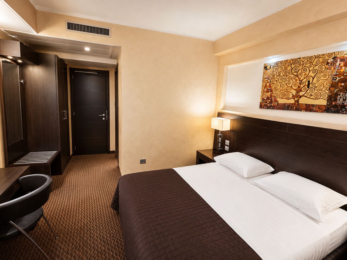 Una habitación de hotel con cama, escritorio, silla, mesita de noche con lámpara y una puerta.