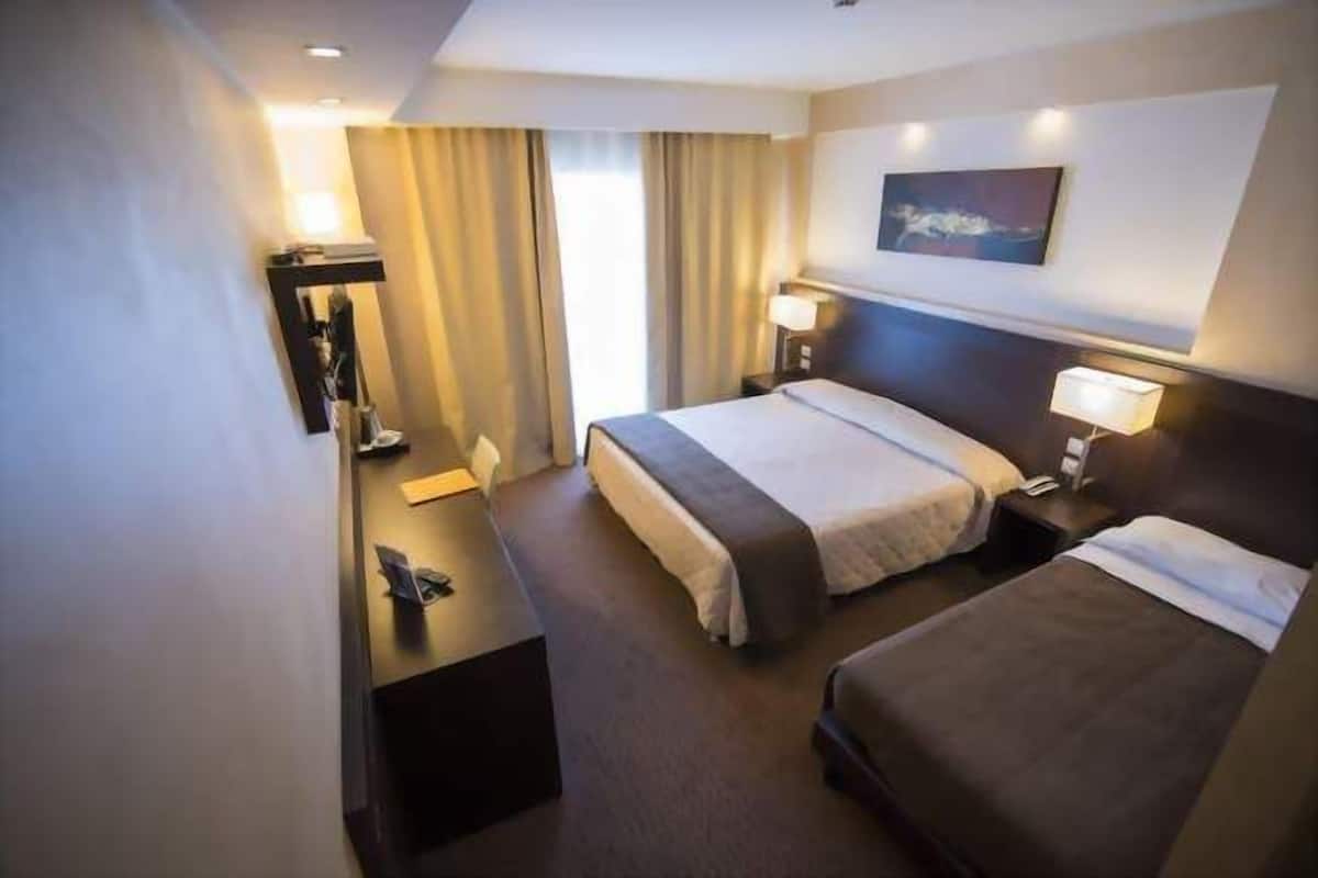A hotel room with two beds, a desk, a chair, a wardrobe, and a painting on the wall.