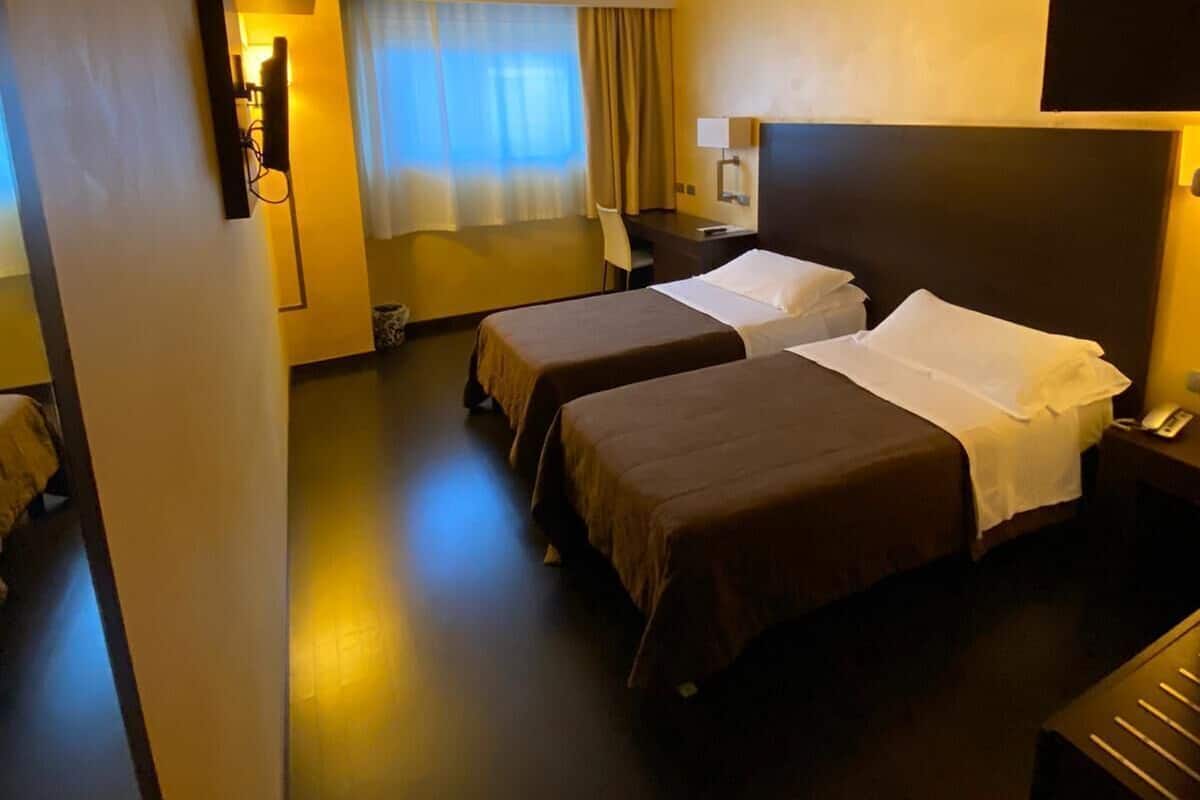 A hotel room with two beds, a desk, a chair, a window with curtains, and a wall-mounted TV.