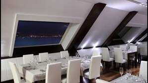 Un espace repas avec des tables dressées pour le service, de grandes fenêtres offrant une vue sur la ville la nuit, et un aménagement intérieur moderne.