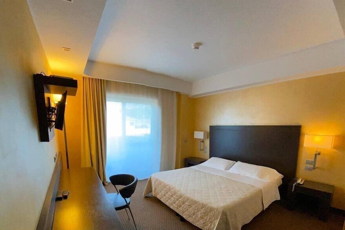 A hotel room with a bed, a desk, a chair, a TV mounted on the wall, and a window with curtains.
