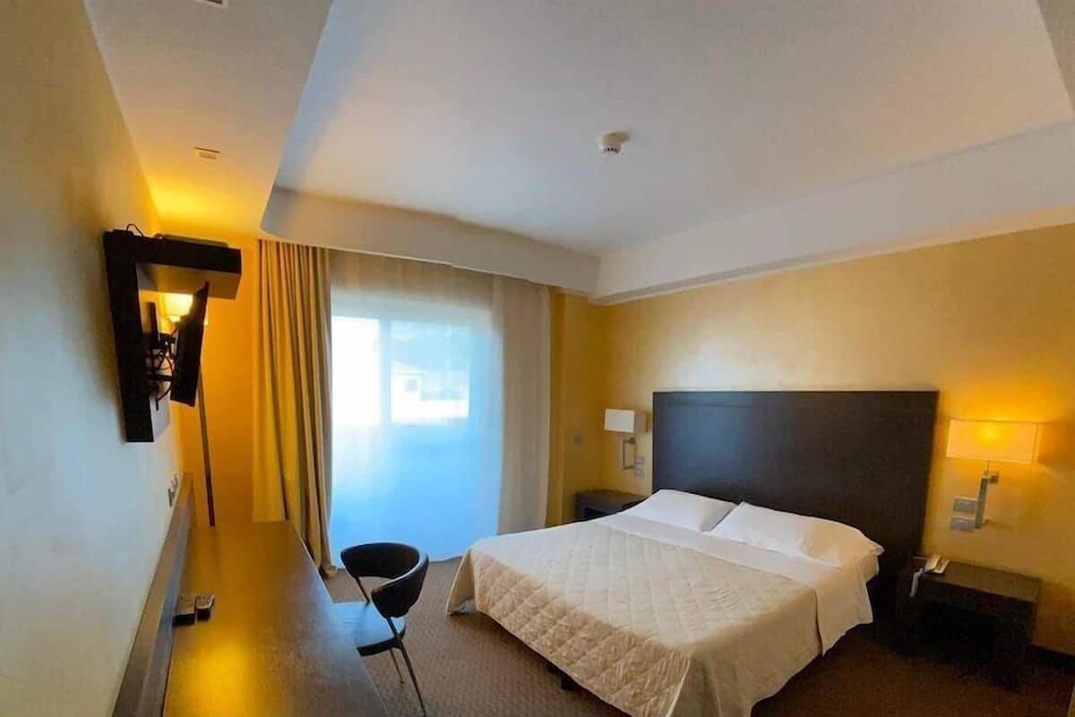 Ein Hotelzimmer mit Bett, Schreibtisch, Stuhl, Wandfernseher und einem Vorhangfenster.
