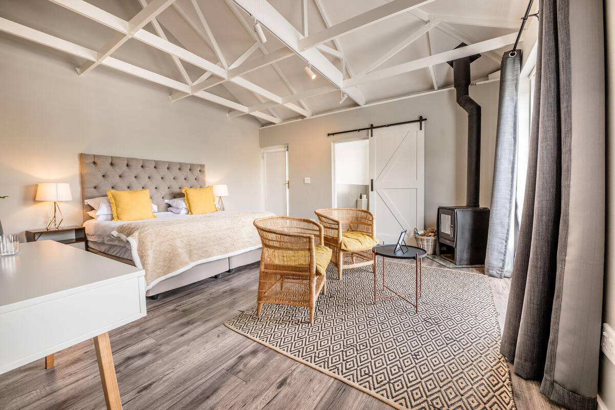 A bedroom with a bed, two chairs, a table, a patterned rug, a ceiling with exposed beams, and a fireplace.