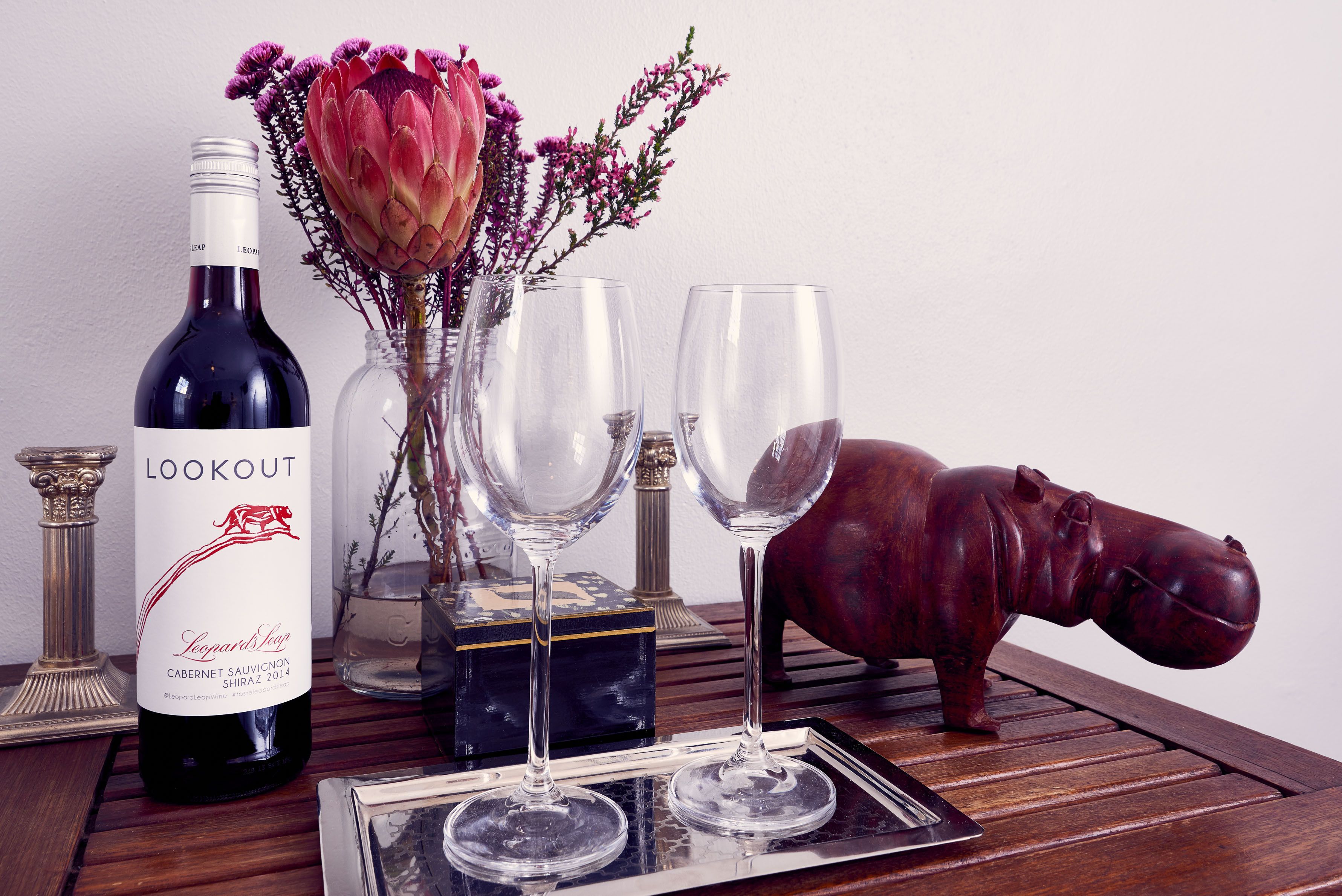 A bottle of Lookout wine, two empty wine glasses, a decorative hippo, and a vase with flowers on a wooden table.