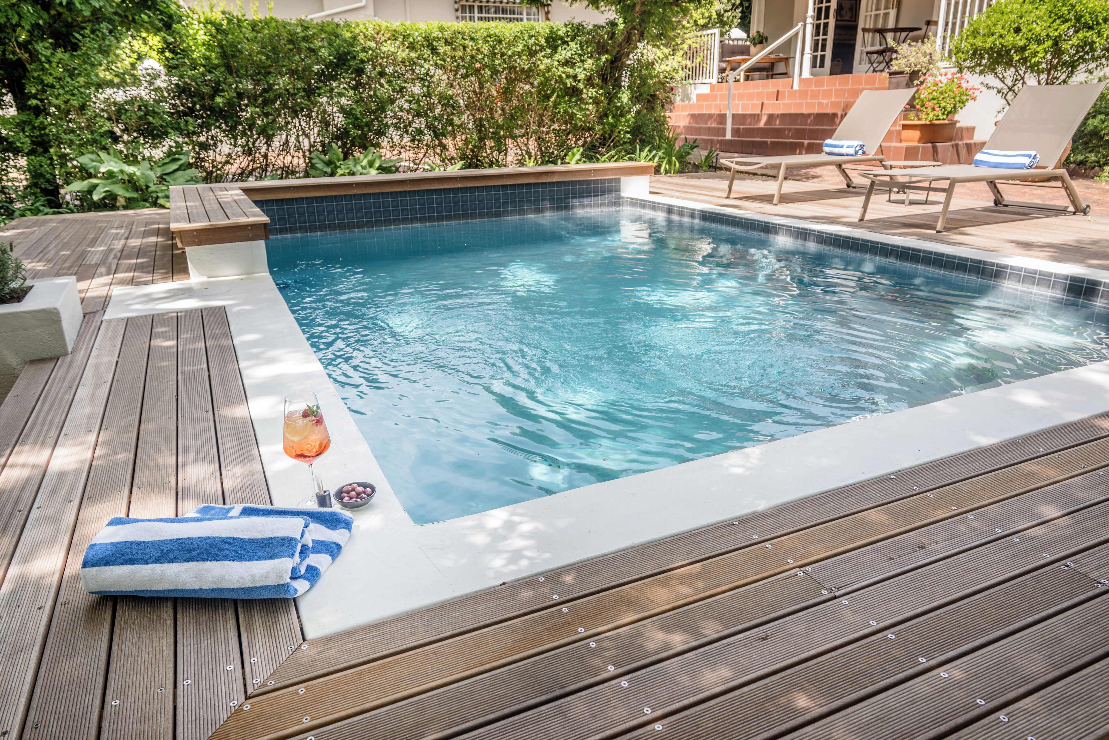 A swimming pool with a wooden deck, lounge chairs, and a drink on the side.