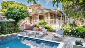Een zwembad met ligbedden, een houten terras en een huis op de achtergrond.