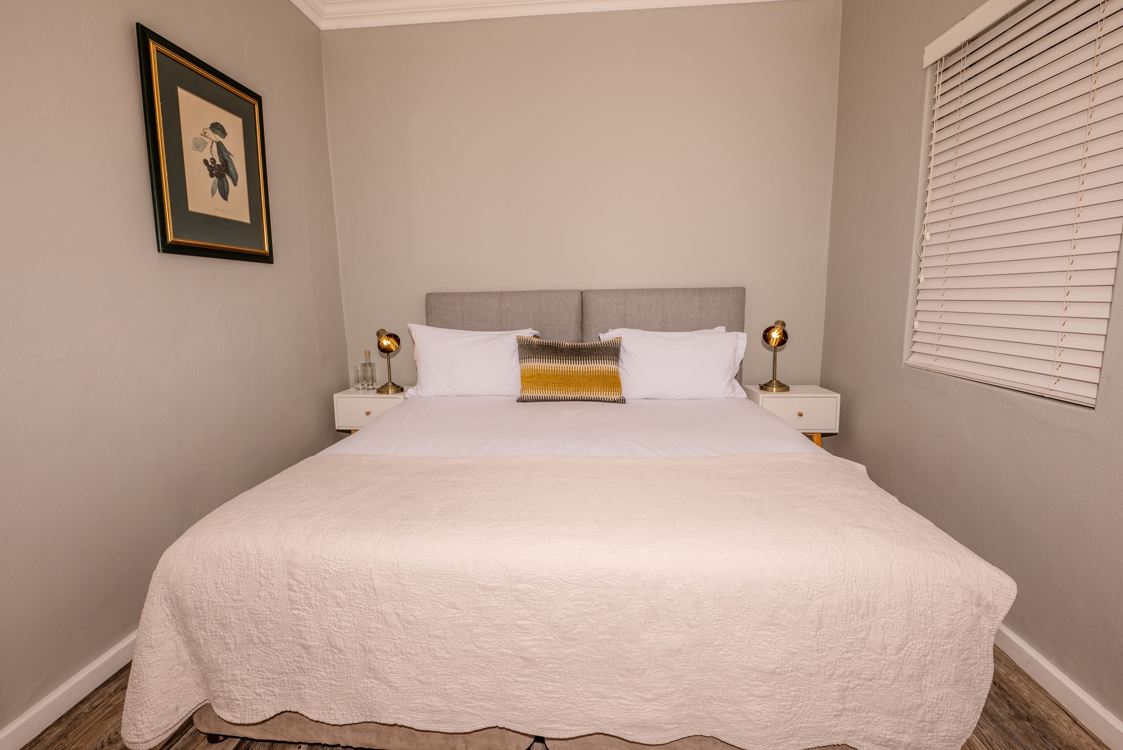 A neatly made bed with a white quilt, two bedside tables with lamps, a framed picture on the wall, and a window with blinds.