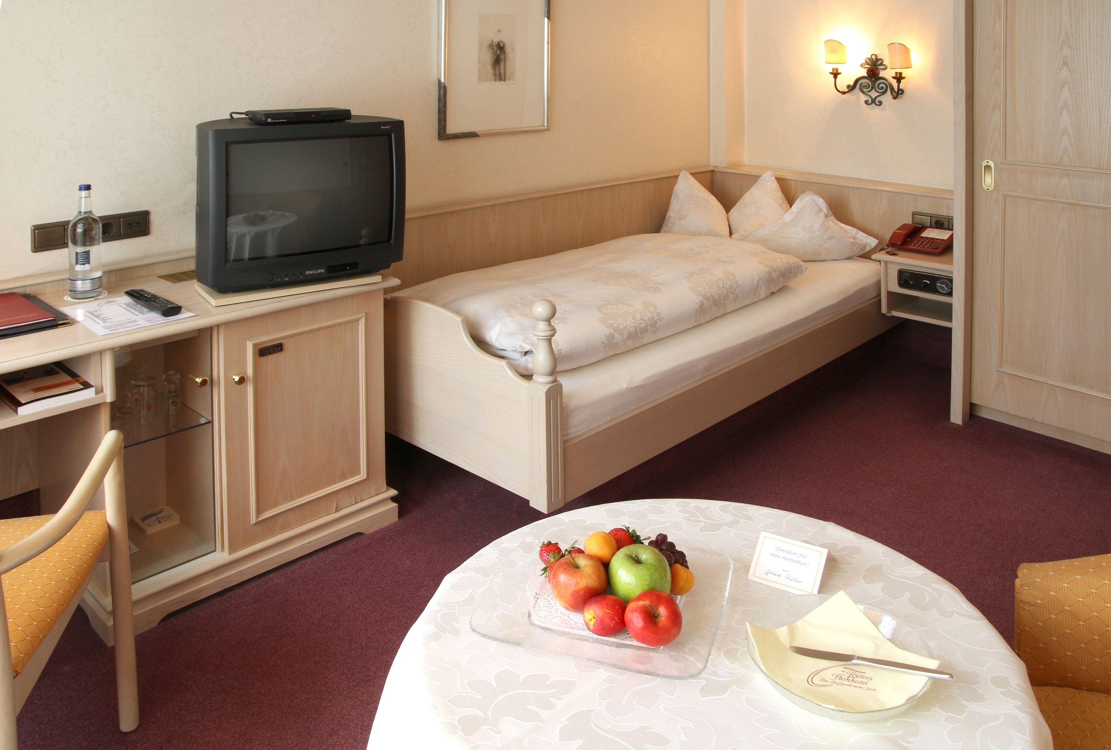 A hotel room with a single bed, a desk with a television, a small table with fruit and a menu, and a chair.