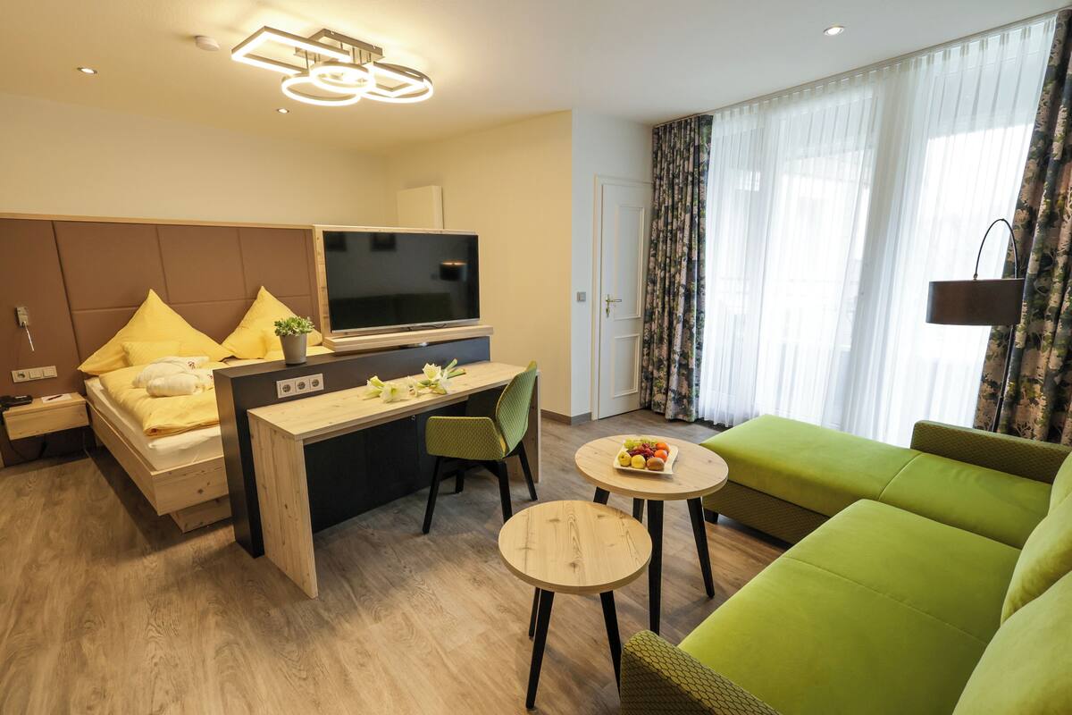 A modern hotel room with a bed, a desk, a television, a green sofa, and a small round table.