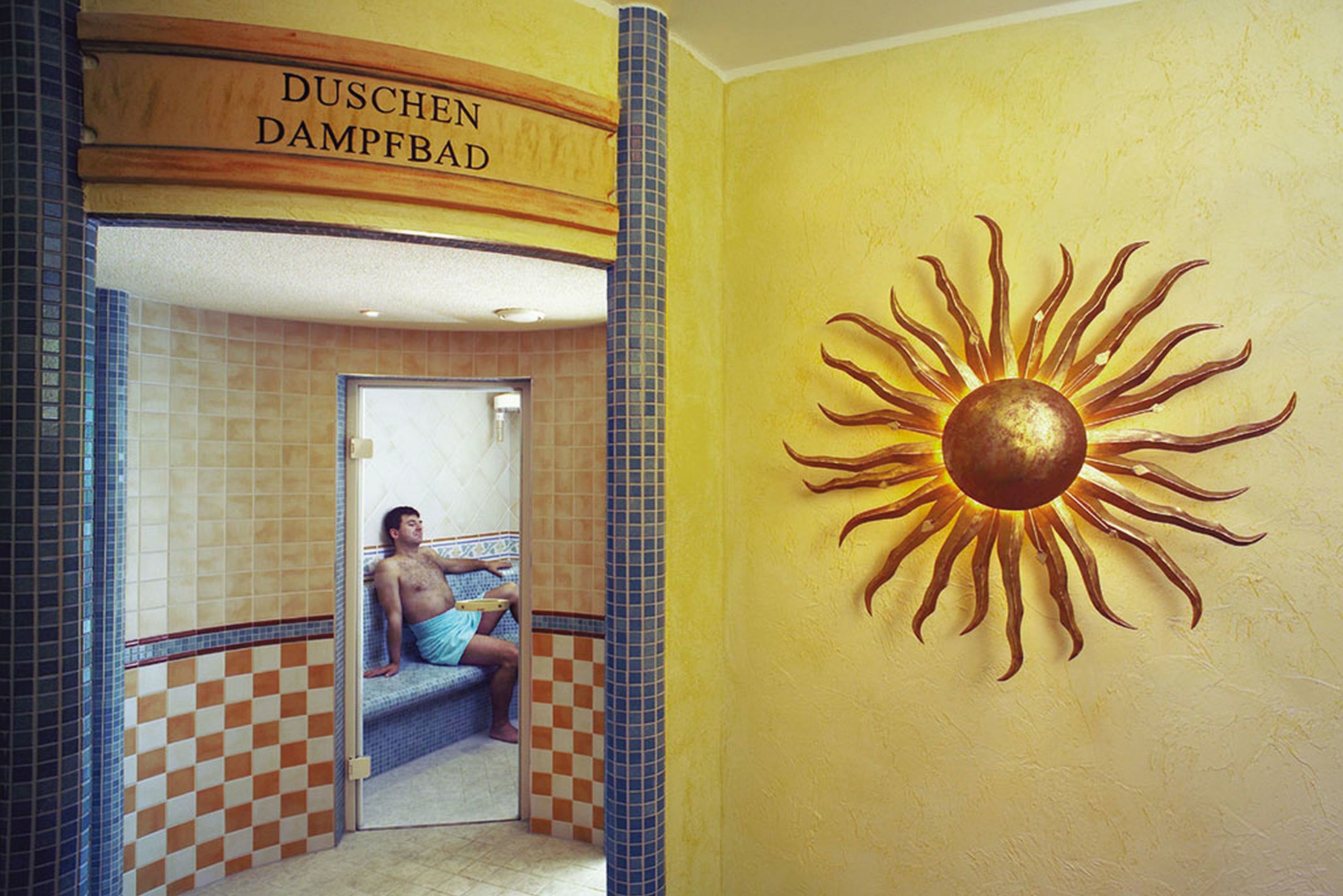 A sun mural and a sign reading "DUSCHEN DAMPFBAD" in a bathroom with checkered tile walls.