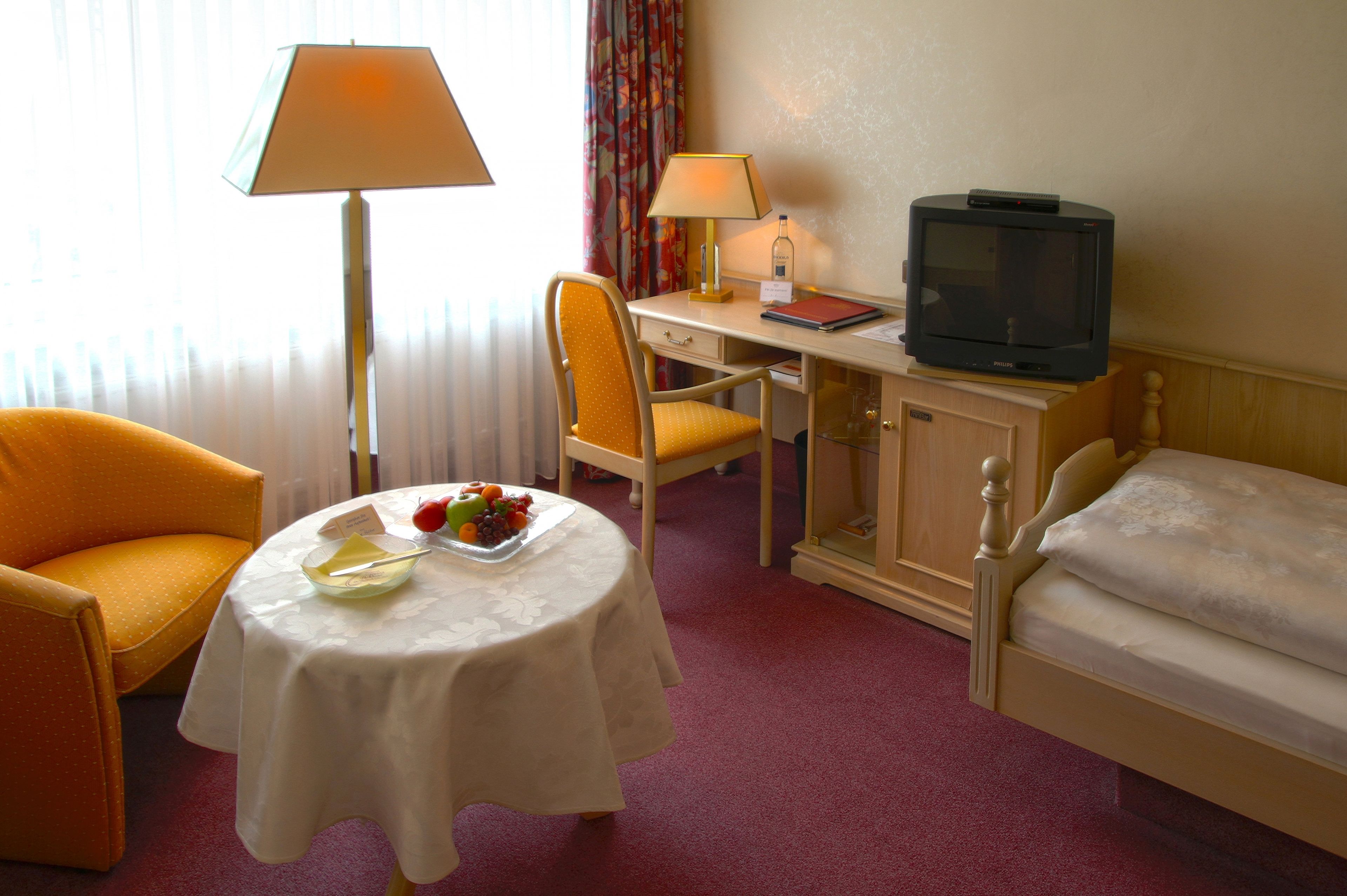 A hotel room with a bed, a desk with a lamp, a television, a chair, and a table with fruit.