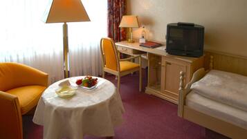 A hotel room with a bed, a desk with a lamp, a television, a chair, and a table with fruit.