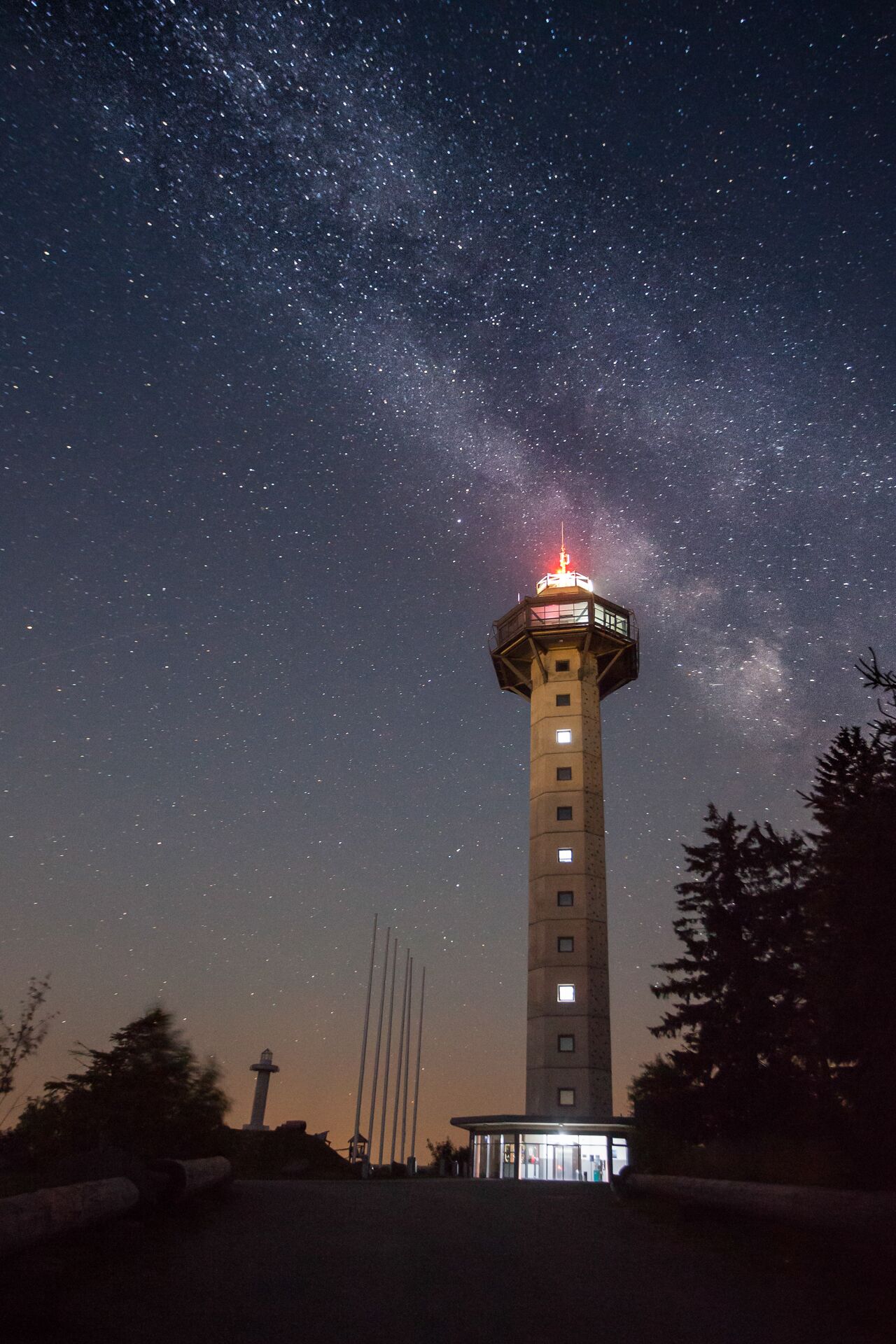 Une haute tour surmontée d’une lumière, entourée d’arbres et s’élevant vers un ciel nocturne dégagé parsemé d’étoiles.
