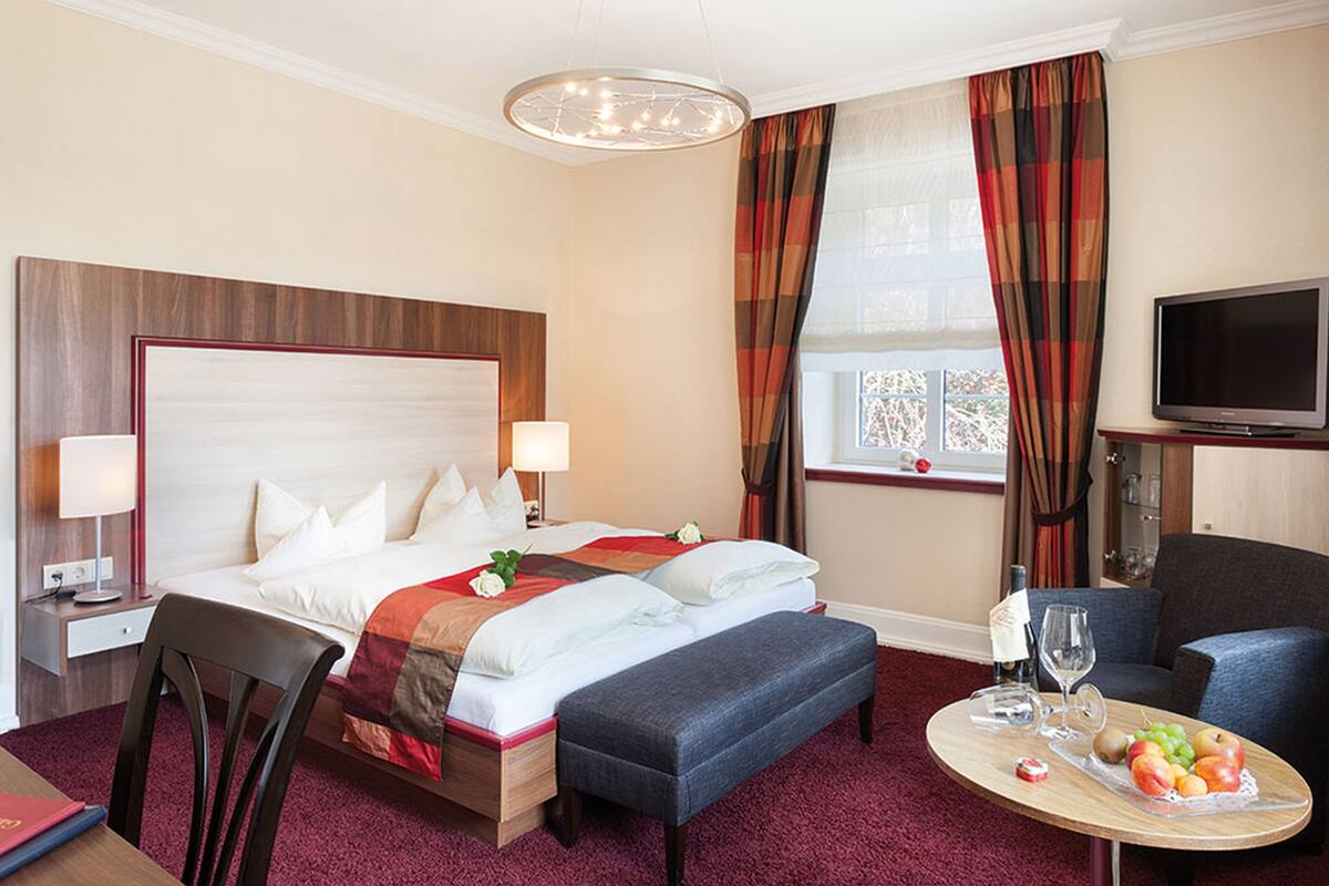 A hotel room with a large bed, a desk with a chair, a small table with a fruit plate, a television, and a window with curtains.