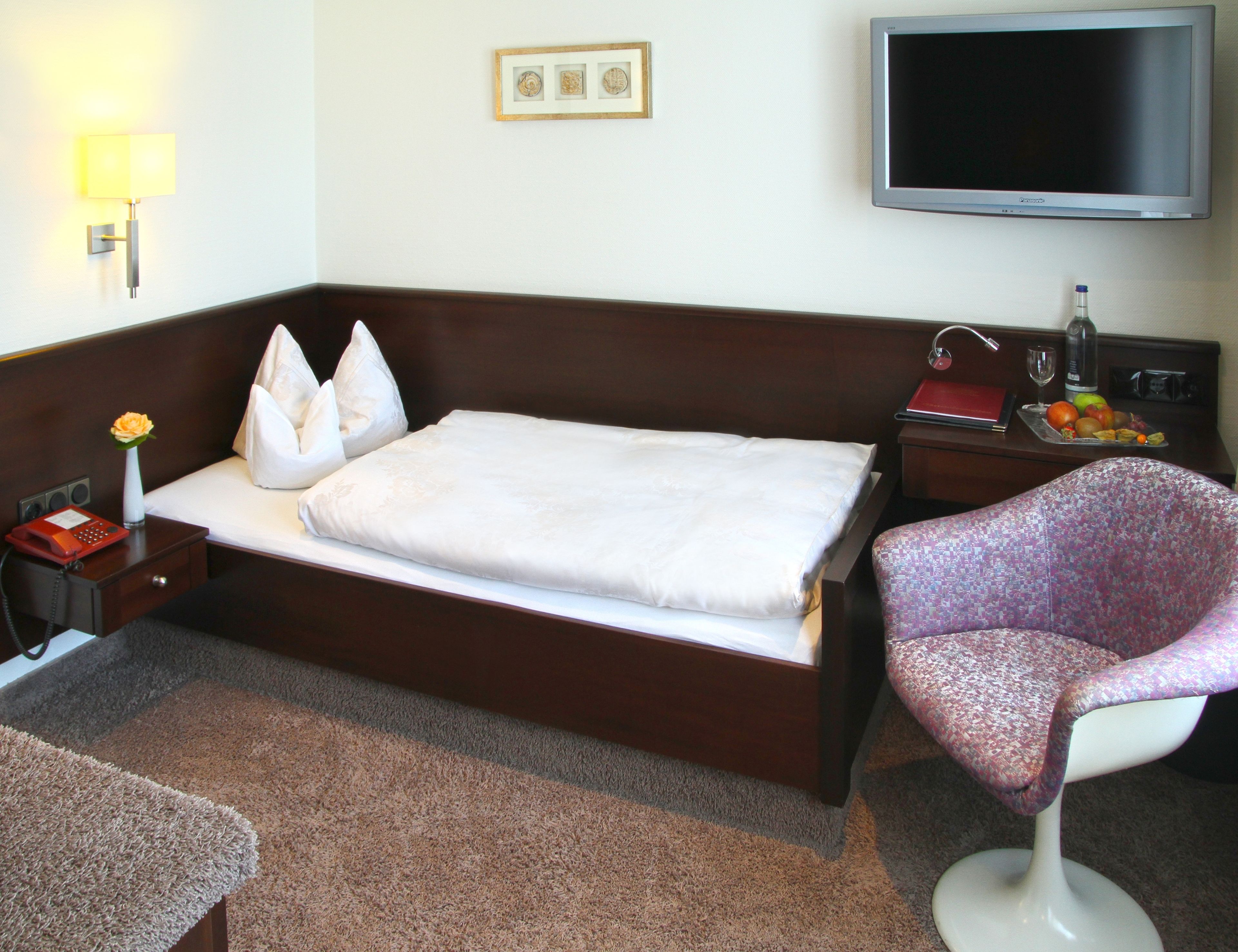 Habitación de hotel con cama, una silla, escritorio con lámpara, televisión y teléfono.
