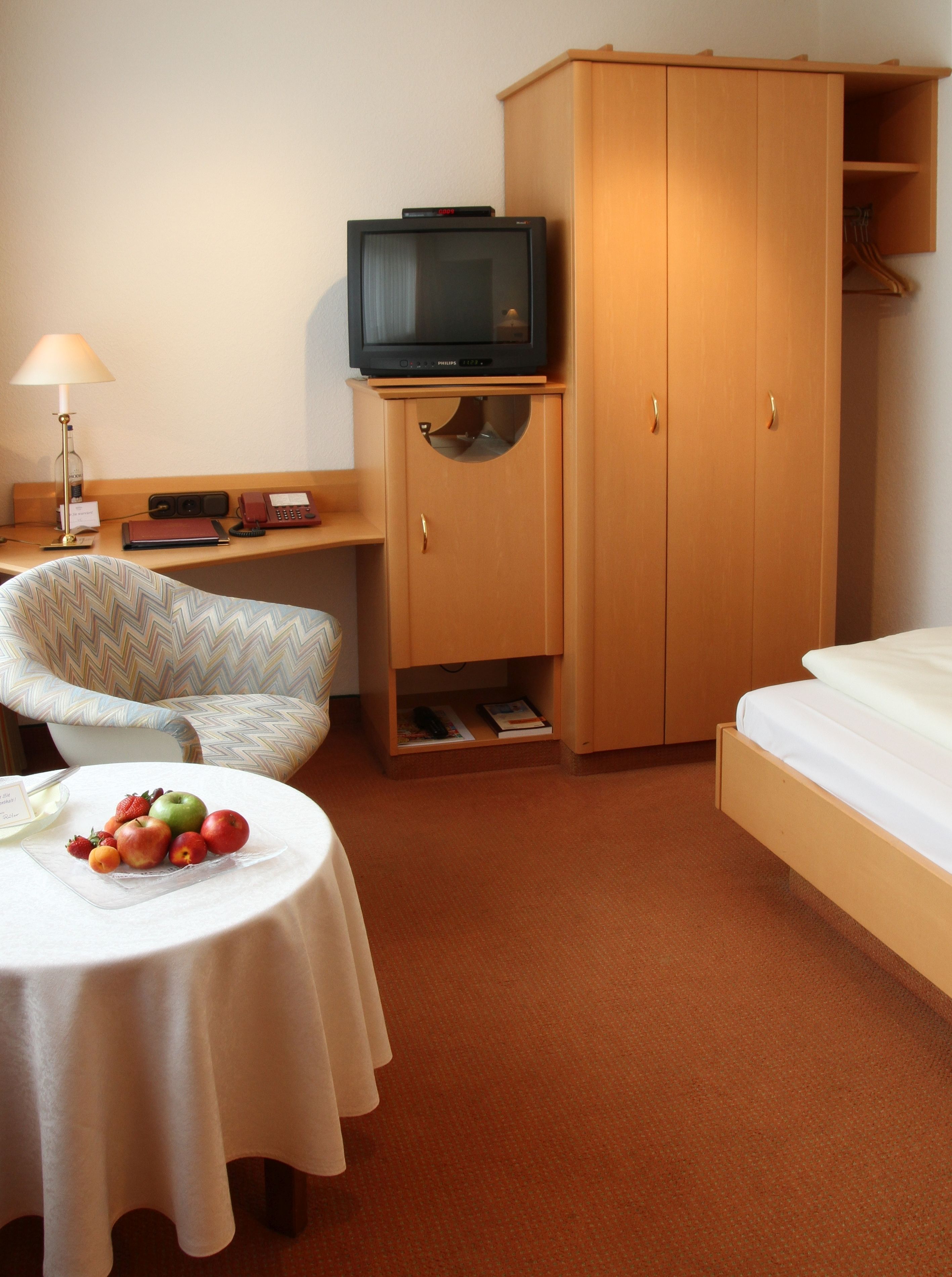 Een hotelkamer met een bed, een televisie op een houten kast, een stoel, een tafel met fruit en een lamp.
