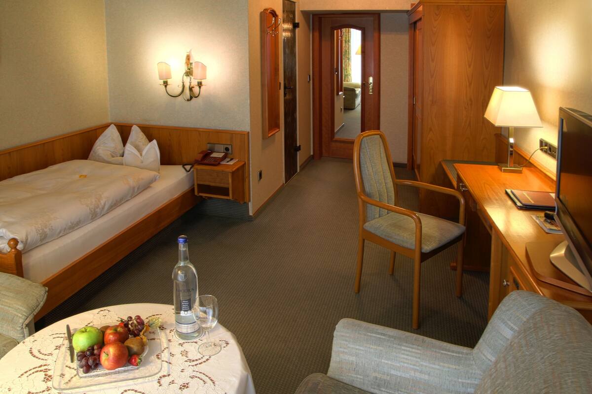 A hotel room with a bed, a desk, a chair, a table with fruit and a bottle, and a mirror.
