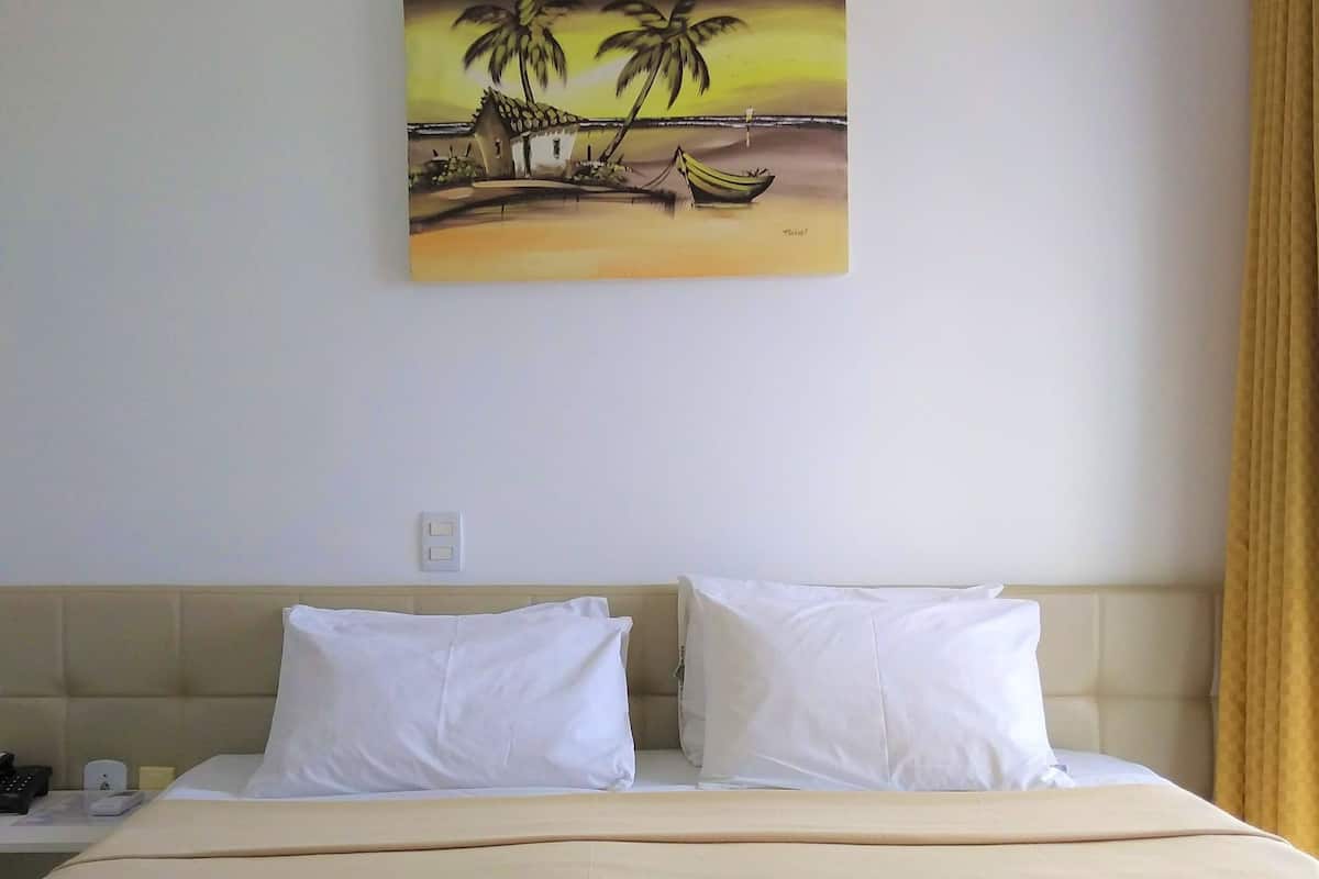 A hotel room with a bed, two pillows, a nightstand, a painting of palm trees and a boat, and a window with curtains.