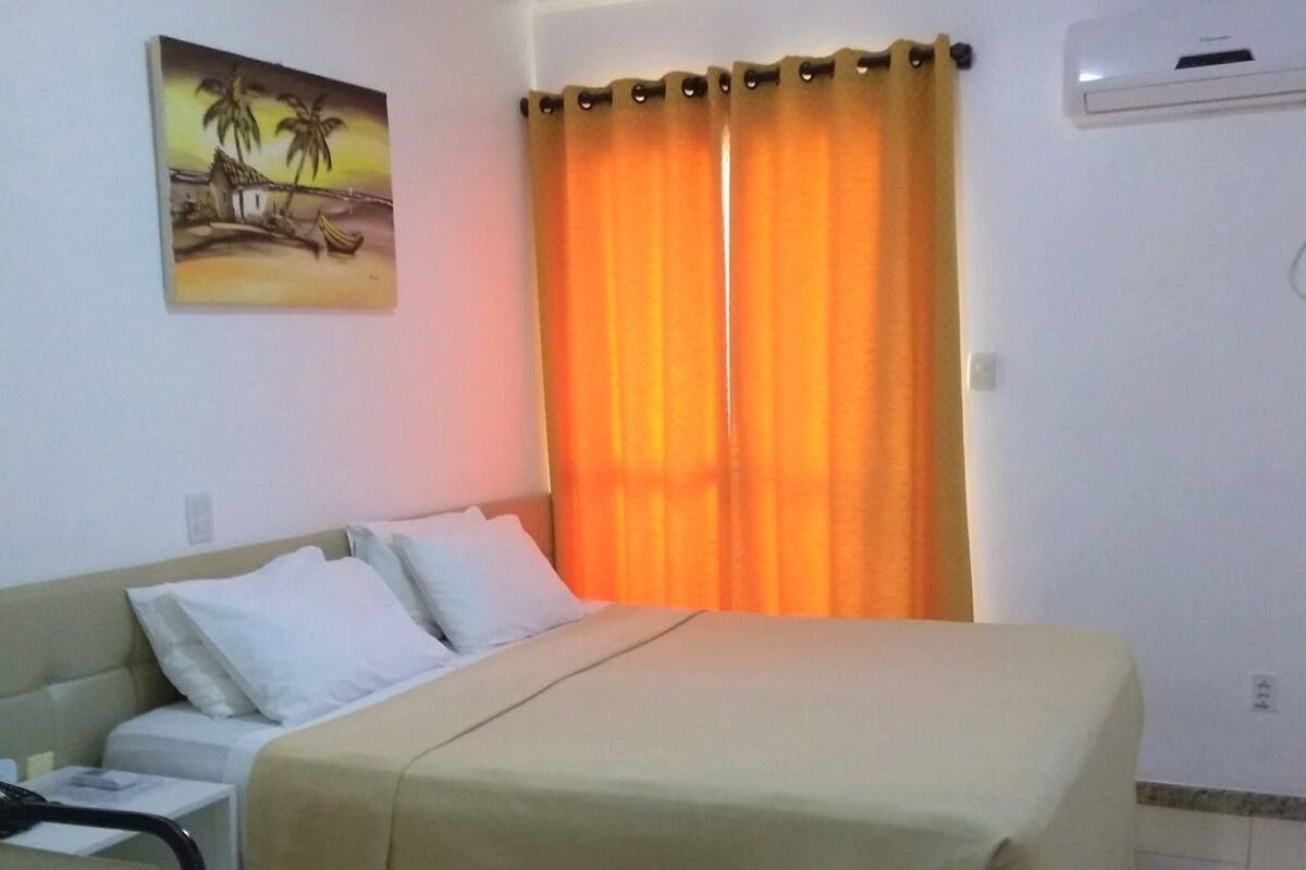 A hotel room with a bed, a bedside table, a chair, a painting on the wall, and an air conditioning unit.