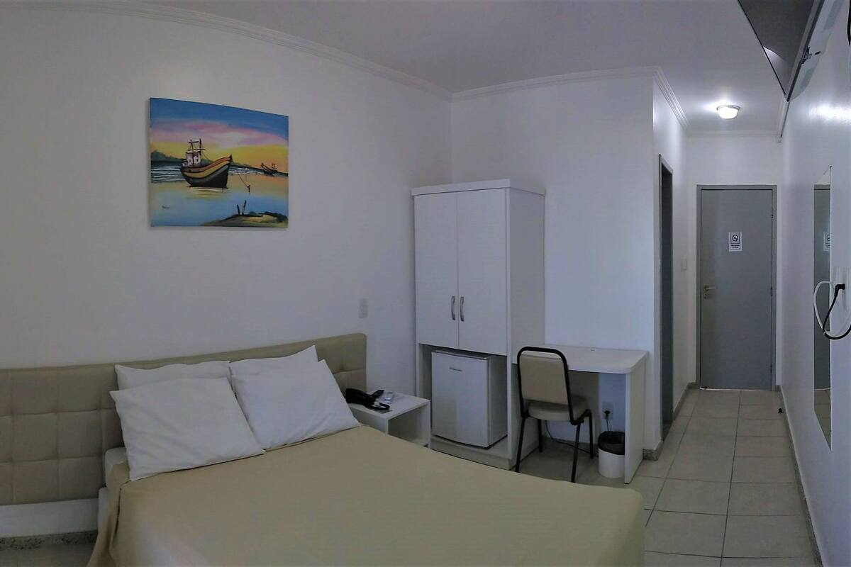 A hotel room with a bed, a desk, a chair, a wardrobe, and a painting of a boat on the wall.