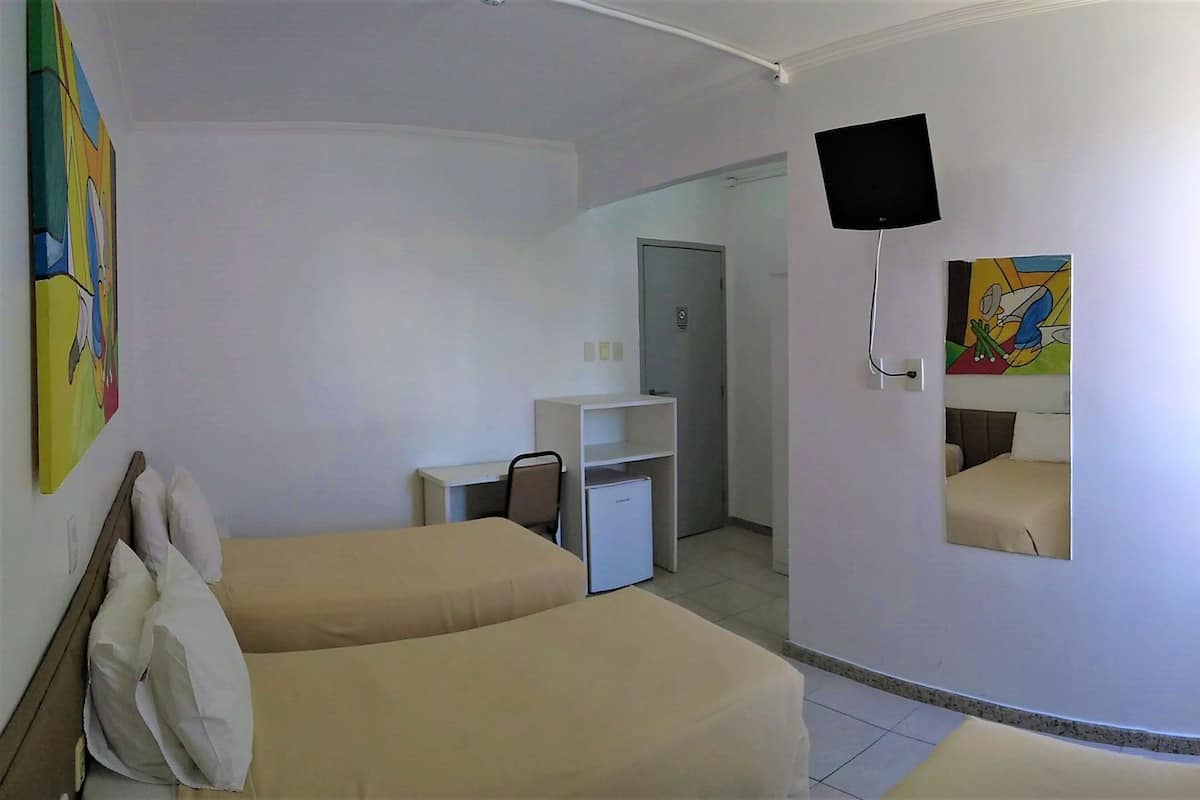 A hotel room with two beds, a small desk, a refrigerator, a TV mounted on the wall, and a door leading to another room.