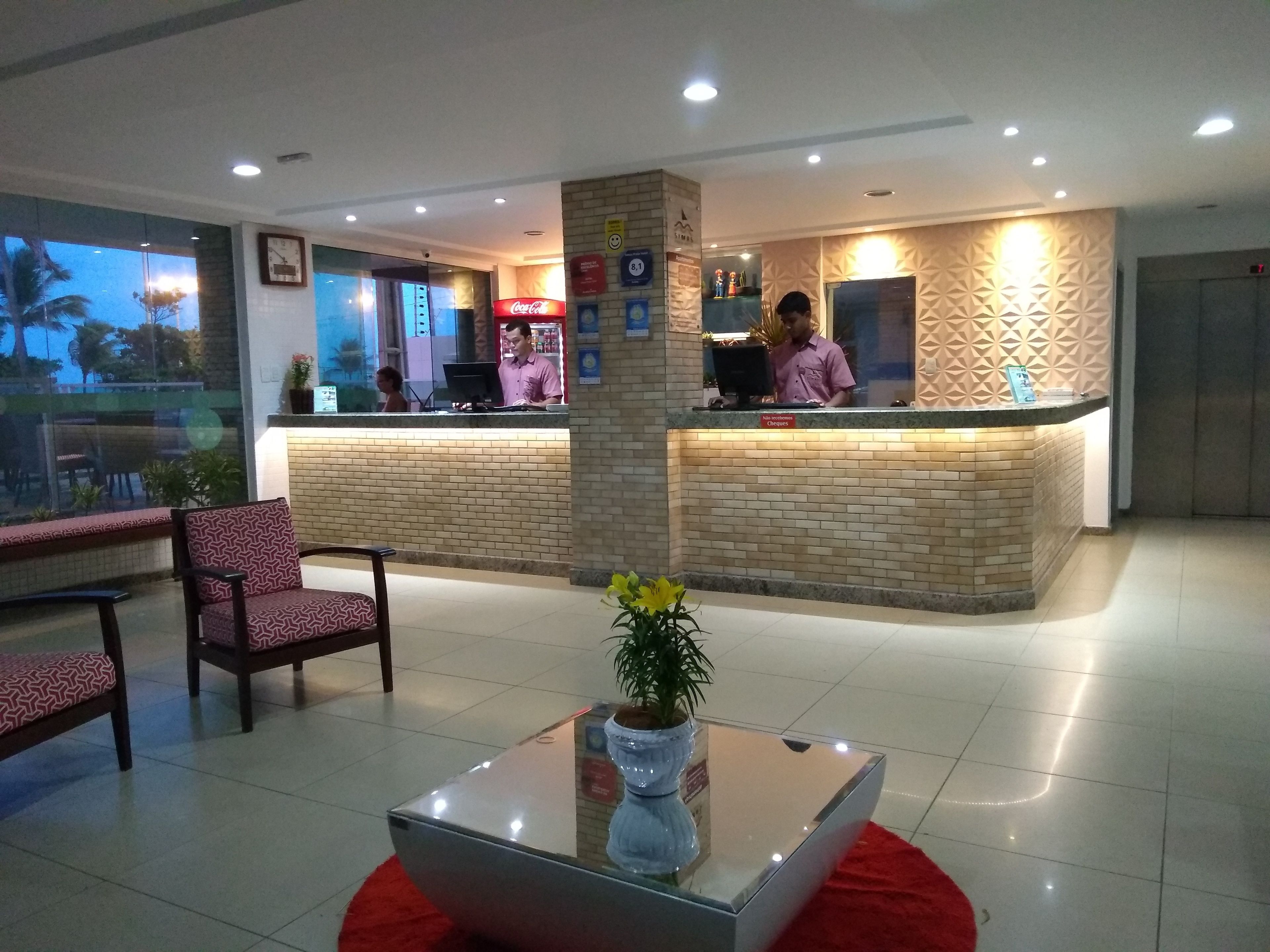 Uma área de recepção de hotel com balcão, computador e relógio na parede. Há cadeiras e uma mesa com uma planta em primeiro plano.