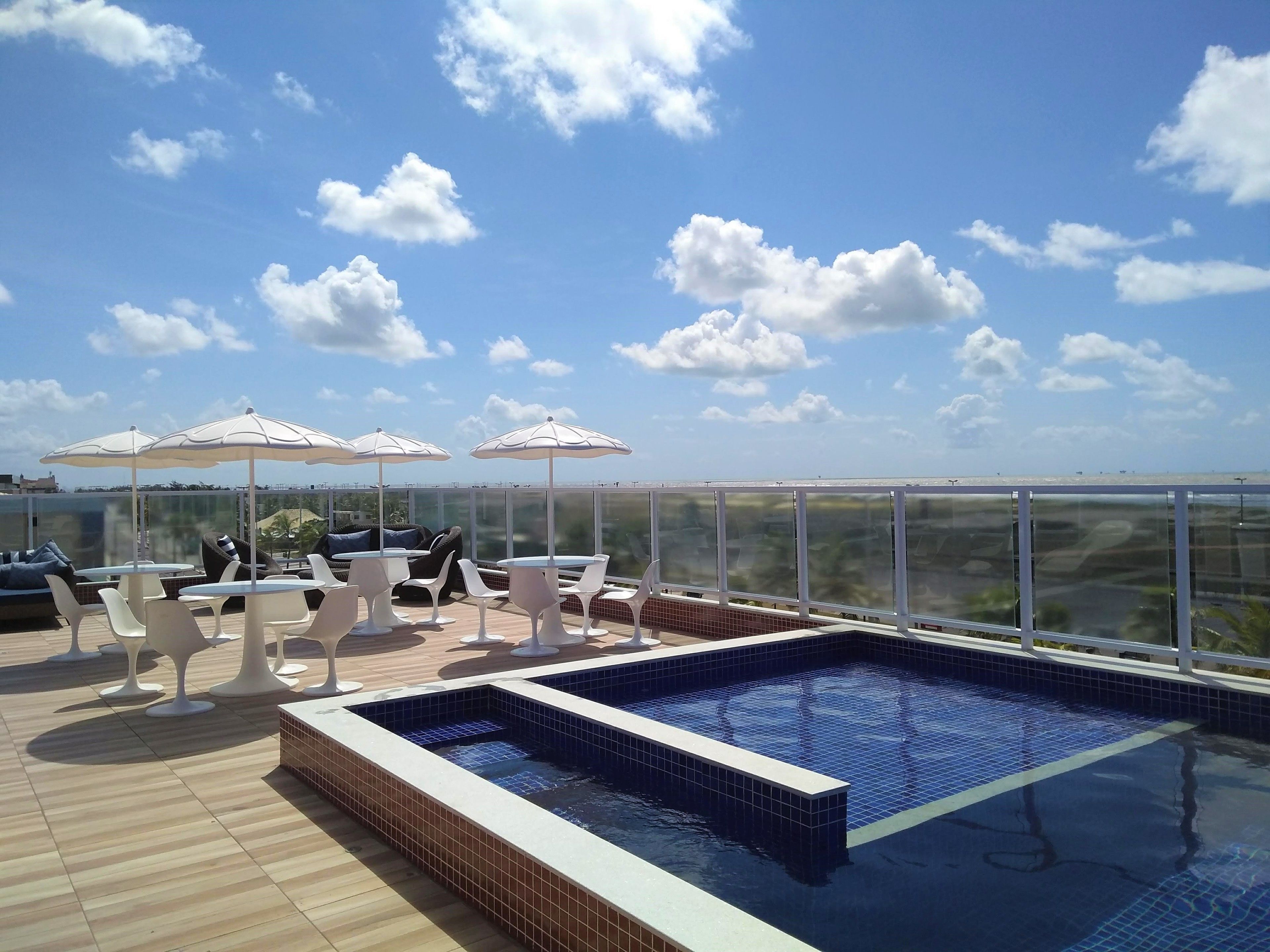 Uma área de piscina no terraço com piso ladrilhado, cadeiras de lounge brancas e guarda-sóis.