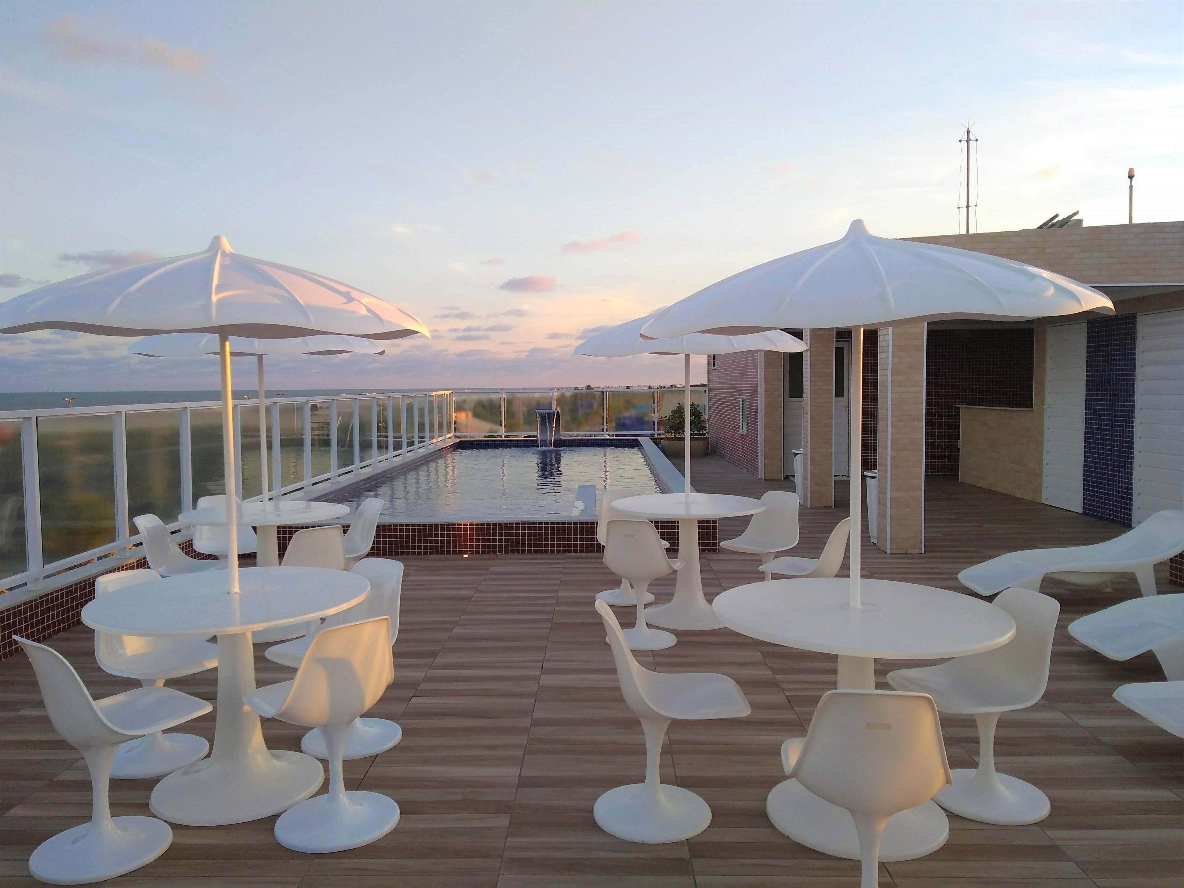 Un área de piscina en la azotea con sillones blancos, mesas, sombrillas y vistas al mar.