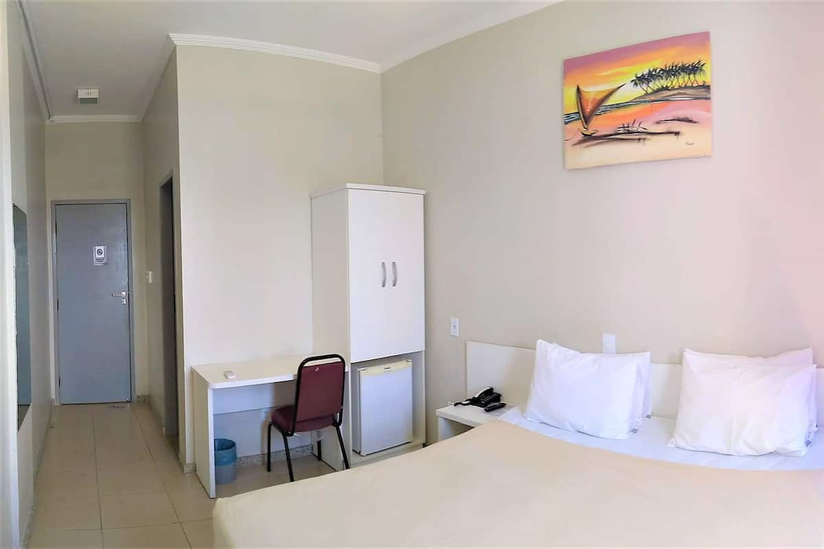 A hotel room with a bed, a desk, a chair, a wardrobe, and a painting on the wall.