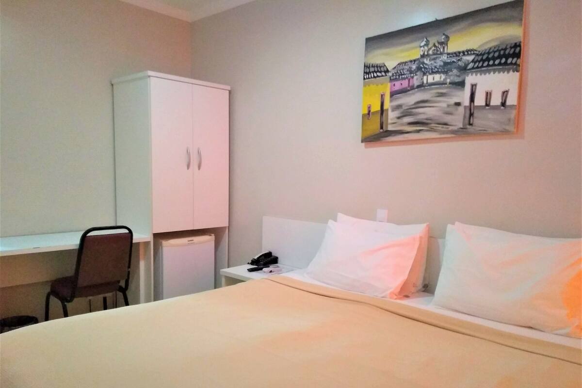 A hotel room with a bed, a desk, a chair, a wardrobe, and a painting on the wall.