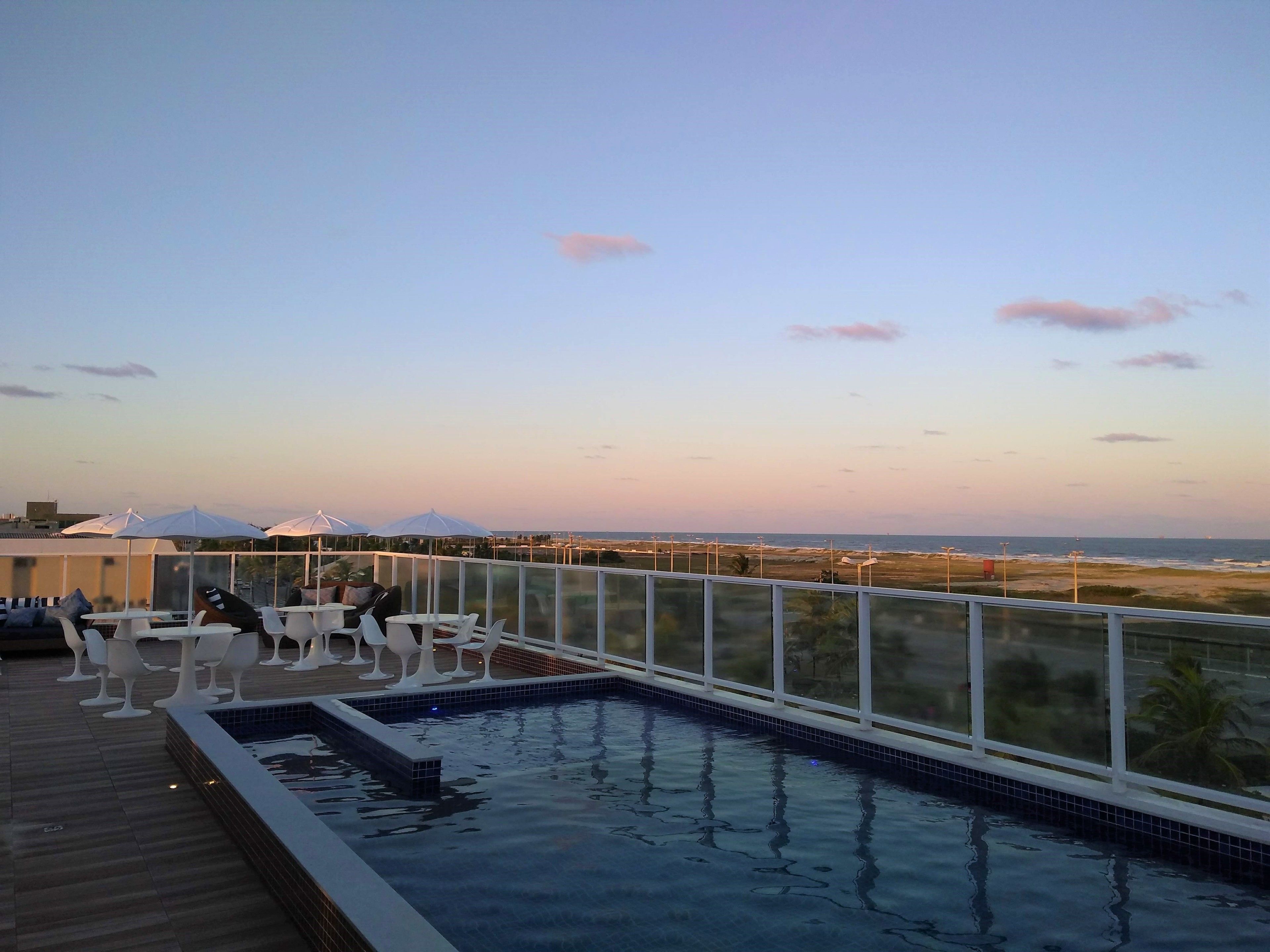 A rooftop pool area with lounge chairs and umbrellas, overlooking a beach and ocean.