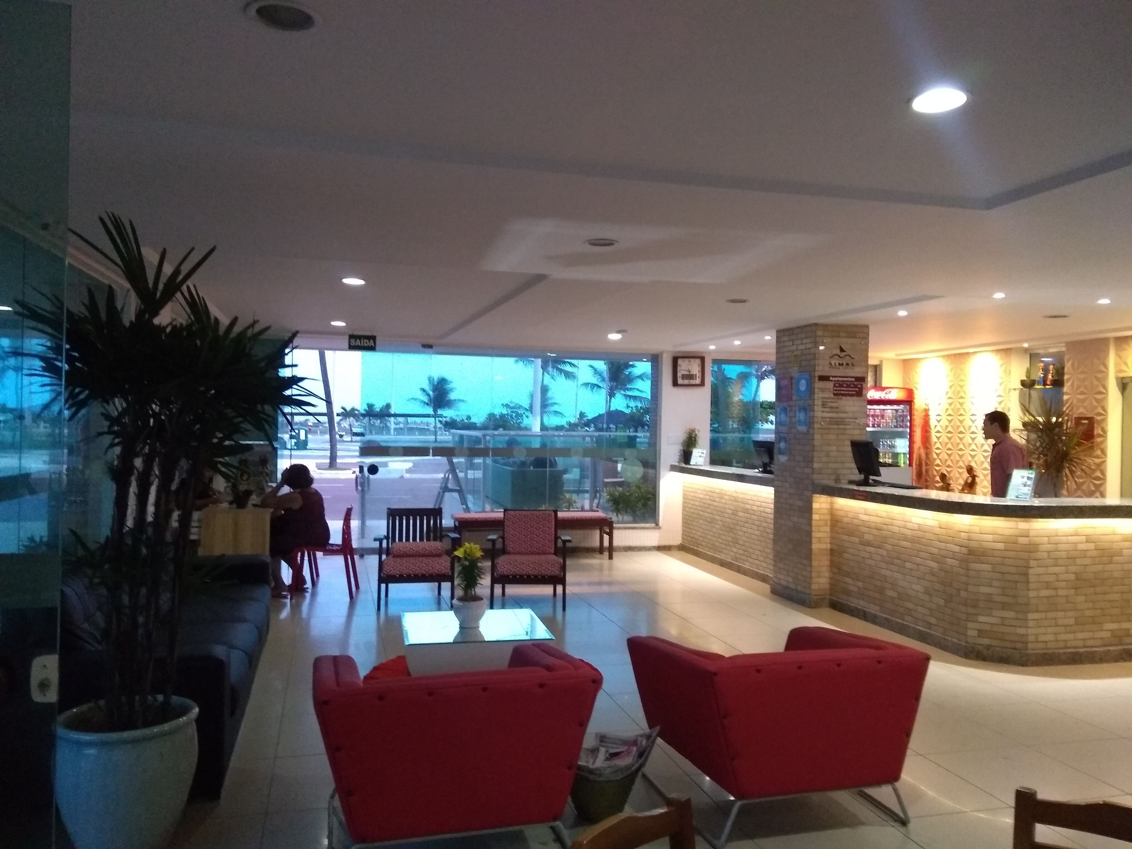 A hotel lobby with red seating, a reception desk, and a view of palm trees outside.