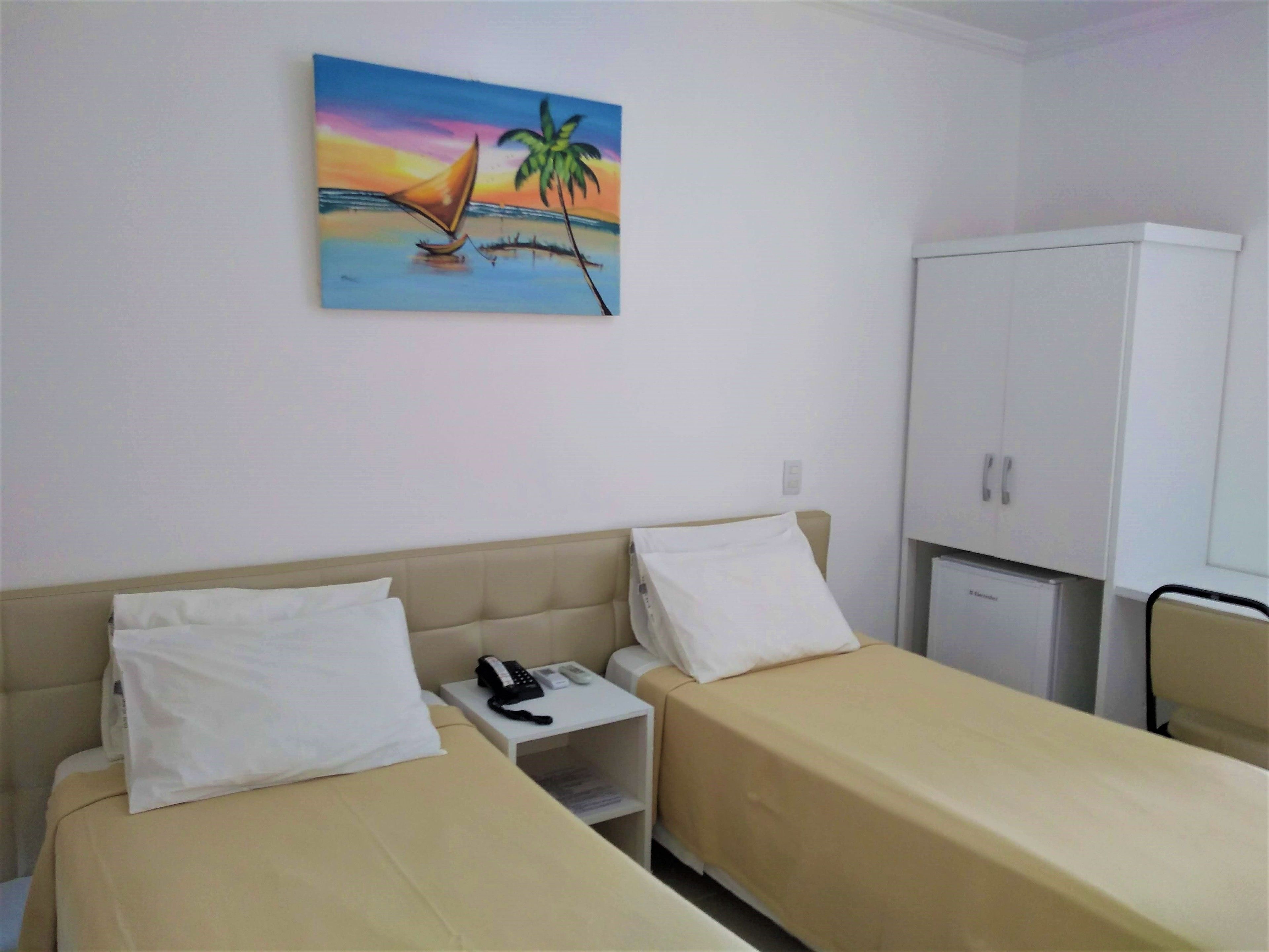 Habitación de hotel con dos camas individuales, una mesita de noche con teléfono, un armario y un cuadro de un velero y una palmera.
