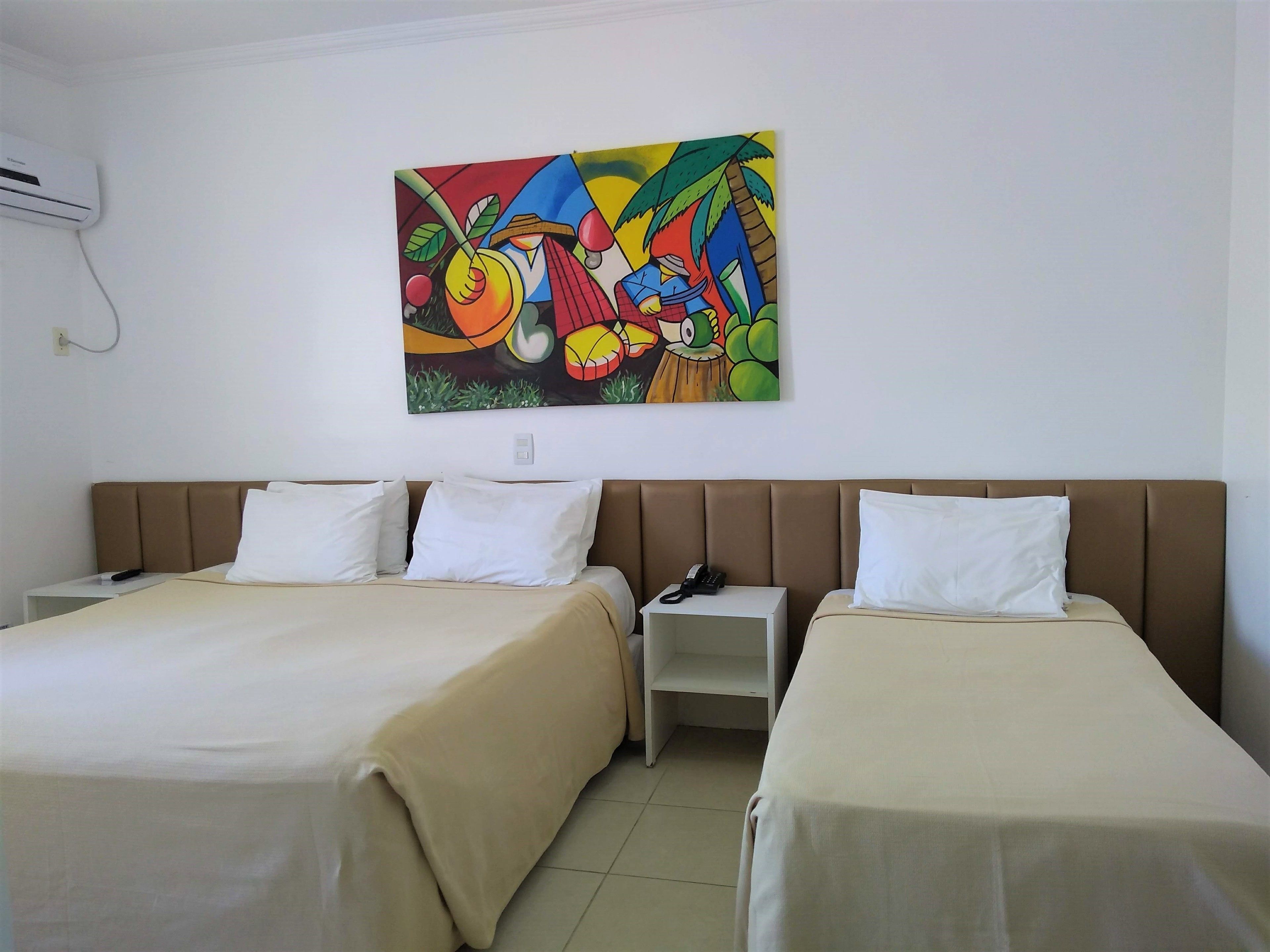 Quarto de hotel com duas camas, uma mesinha de cabeceira, um telefone e um quadro abstrato colorido na parede.