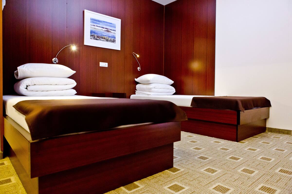 A hotel room with two beds, a wooden headboard, a bedside table, a wall-mounted lamp, and a framed picture on the wall.